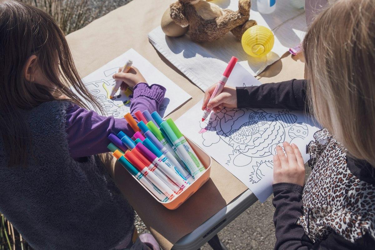 Deux fillettes penchées sur des coloriages à une table, avec une boîte orange remplie de feutres de couleur