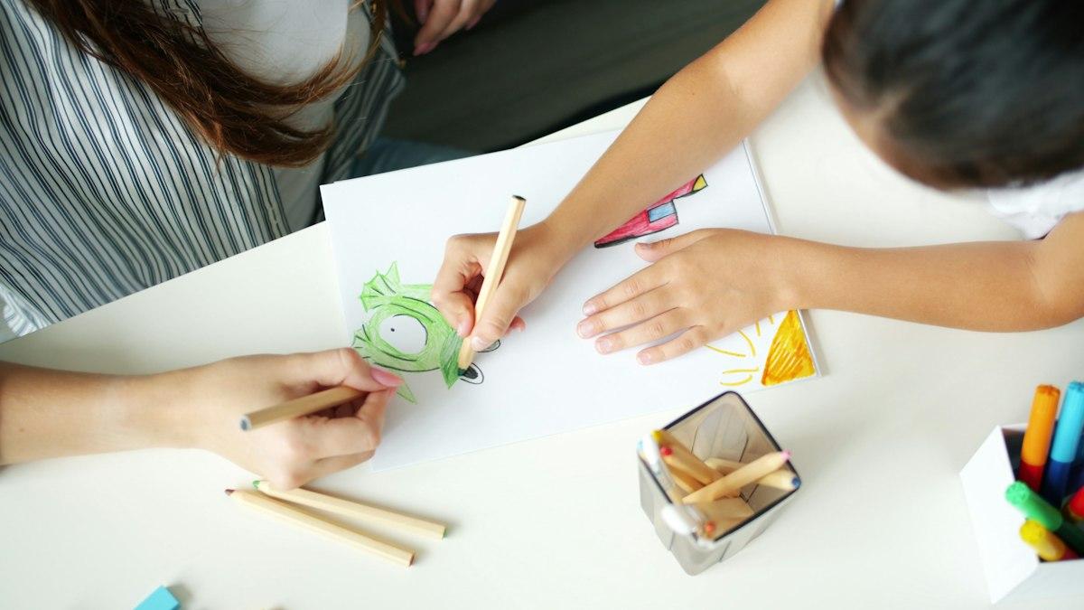 Un adulte et un enfant dessinent côte à côte sur une feuille blanche, crayons de couleur posés sur la table